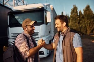 Happy truck driver and his coworker greeting while standing on parking lot.